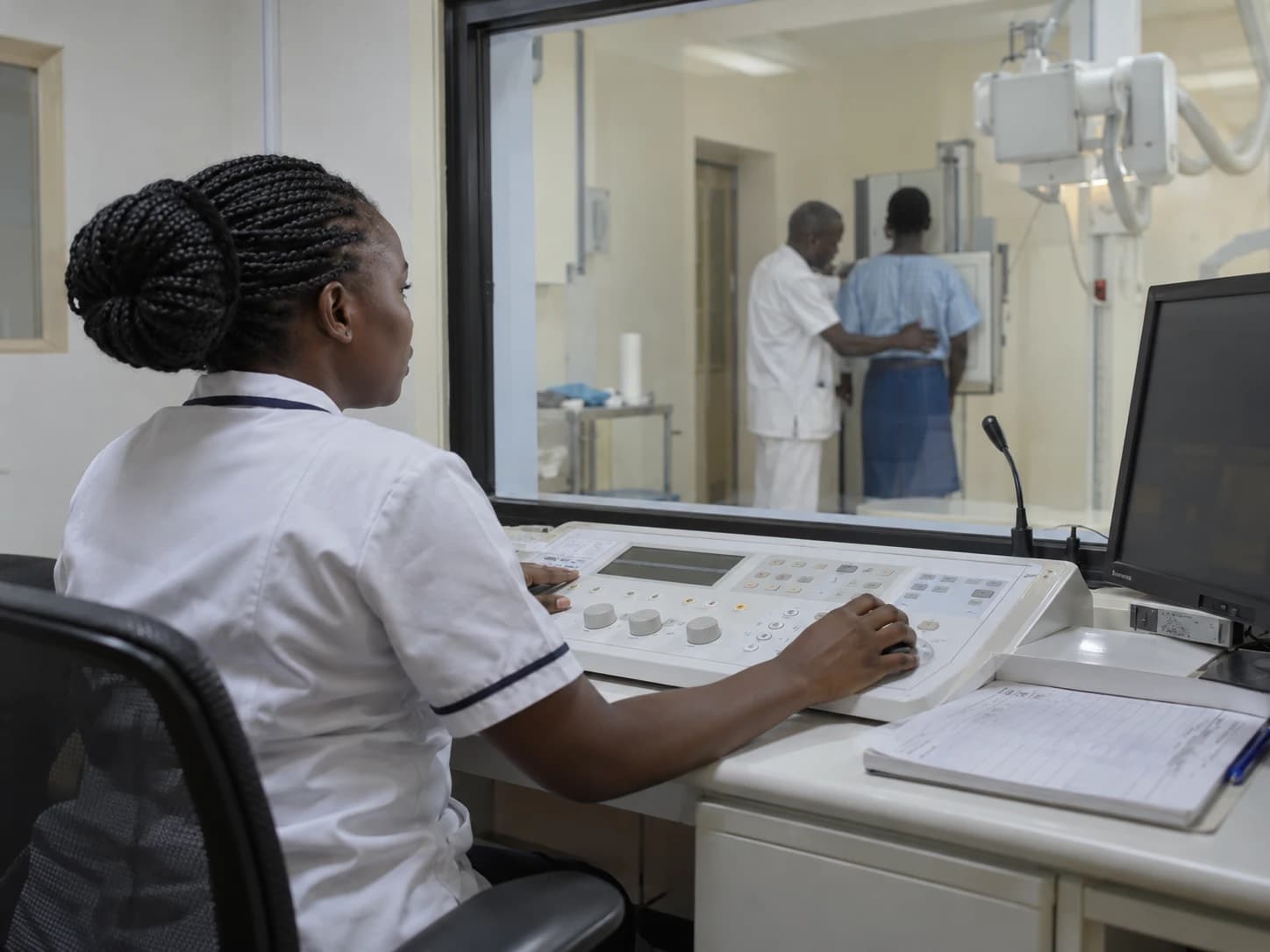 A radiographer at an X-ray control console looking through the viewing window